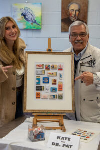 Kate Malooley, left, and Dr. Pat Polis, right, standing indoors and smiling while pointing at a framed artwork made of arranged matchbooks on an easel, with paintings displayed on the wall behind them.