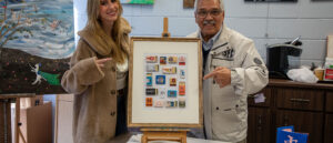 Kate Malooley, left, and Dr. Pat Polis, right, standing indoors and smiling while pointing at a framed artwork made of arranged matchbooks on an easel, with paintings displayed on the wall behind them.