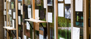 Exhibition installation showing a series of architectural drawings, photographs, and text panels mounted on vertical wire grid panels, with small white architectural models displayed on narrow shelves in front.