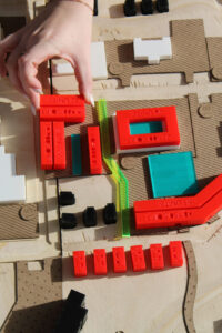 Overhead view of a student urban planning model made of layered wood, with white and black building massing blocks and bright red 3D-printed structures labeled with unit counts. A hand places a red rectangular piece onto the site near a translucent green strip, suggesting building placement within the proposed development.