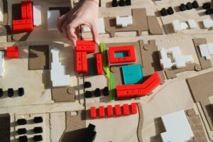 Overhead view of a student urban planning model made of layered wood, with white and black building massing blocks and bright red 3D-printed structures labeled with unit counts. A hand places a red rectangular piece onto the site near a translucent green strip, suggesting building placement within the proposed development.