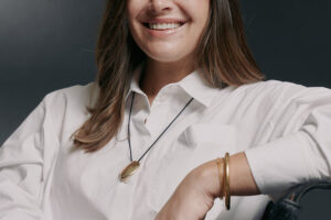 Dani Miller poses for a headshot from the waist up. She sits in a chair with her left arm propped up on her chair.