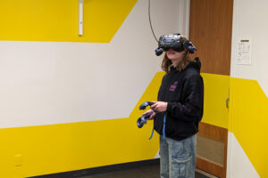 A student wearing a virtual reality headset and holding VR controllers stands in the Digital Futures Lab, immersed in an interactive experience against a bright yellow-and-white geometric wall.
