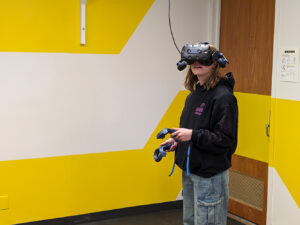 A student wearing a virtual reality headset and holding VR controllers stands in the Digital Futures Lab, immersed in an interactive experience against a bright yellow-and-white geometric wall.