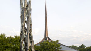 A tall, woven lattice sculpture made of translucent mesh-like tubes rises from a grassy lawn, with a modern church featuring a steep copper-colored spire in the background. Trees surround the site under a pale morning sky.