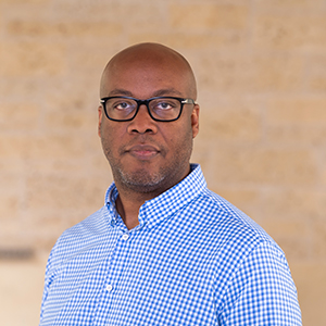 Headshot of a man with a shaved head and glasses, wearing a blue-and-white checkered button-down shirt.