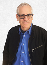 Headshot of a man with short gray hair and glasses, wearing a blue button-down shirt and black jacket.
